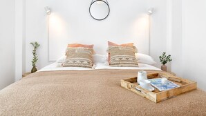 Una cama bien tendida con un edredón tejido, dos almohadones decorativos, una bandeja de madera con tazas y un libro, y luces fijadas a la pared.