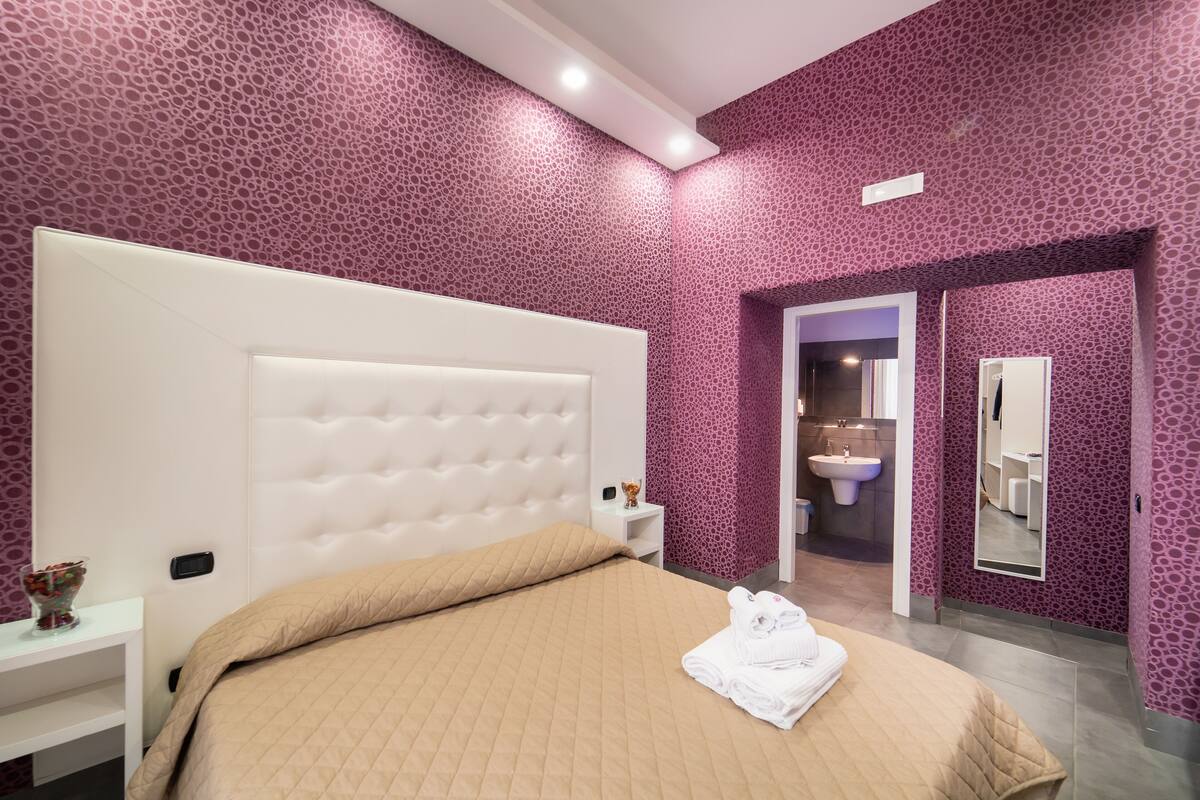 Quarto de hotel com uma cama grande, uma mesinha de cabeceira e um banheiro visível por uma porta aberta.