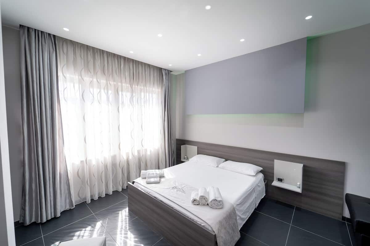 Quarto de hotel moderno com uma cama grande, mesa de cabeceira, televisão fixada na parede e janela com cortinas translúcidas.