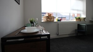 A room with a dining table set for two, a window with a view of buildings, a radiator, and a small chair. - Apartments St Thomas Road (Chorley)