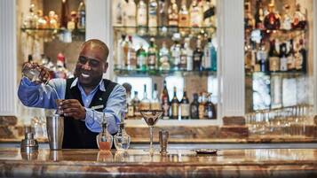 A bar area with a bartender preparing a drink, a variety of bottles and glasses on shelves, and a polished marble countertop.
