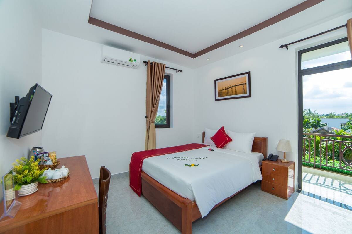 A hotel room with a bed, a desk with a TV, a window with a view, and a balcony with a railing.