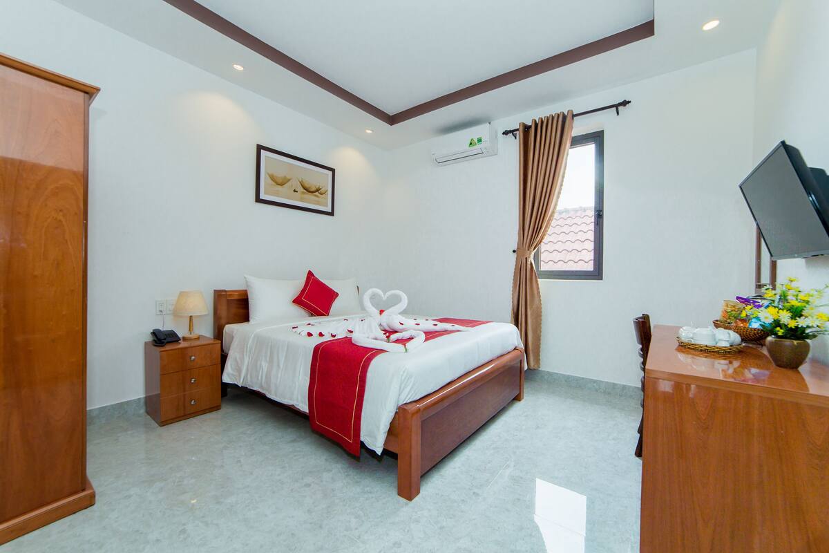 A hotel room with a bed, a wooden nightstand, a desk, a television, and a window with curtains.