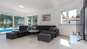 Ein modernes Wohnzimmer mit einem Ecksofa, Hocker, einem Couchtisch und einem Flachbildfernseher. Große Fenster bieten einen Blick ins Freie.