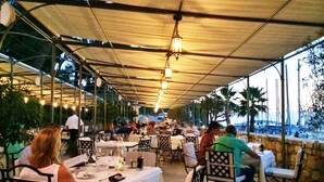 Un espace de restauration en plein air, avec des tables et des chaises disposées sous une pergola, donnant sur un port de plaisance.