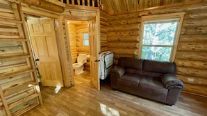 Interno di una baita di legno con pavimento in legno, un divano in pelle marrone, una finestra con vista sugli alberi e una porta che conduce al bagno.