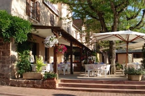 An outdoor café with tables and chairs, a covered seating area, and a building with a stone facade. - Auberge Aux Portes de Conques (Conques-en-Rouergue)