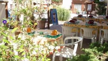 Un área de comedor al aire libre con sillas y mesas de plástico blanco, rodeada de follaje y plantas en macetas.