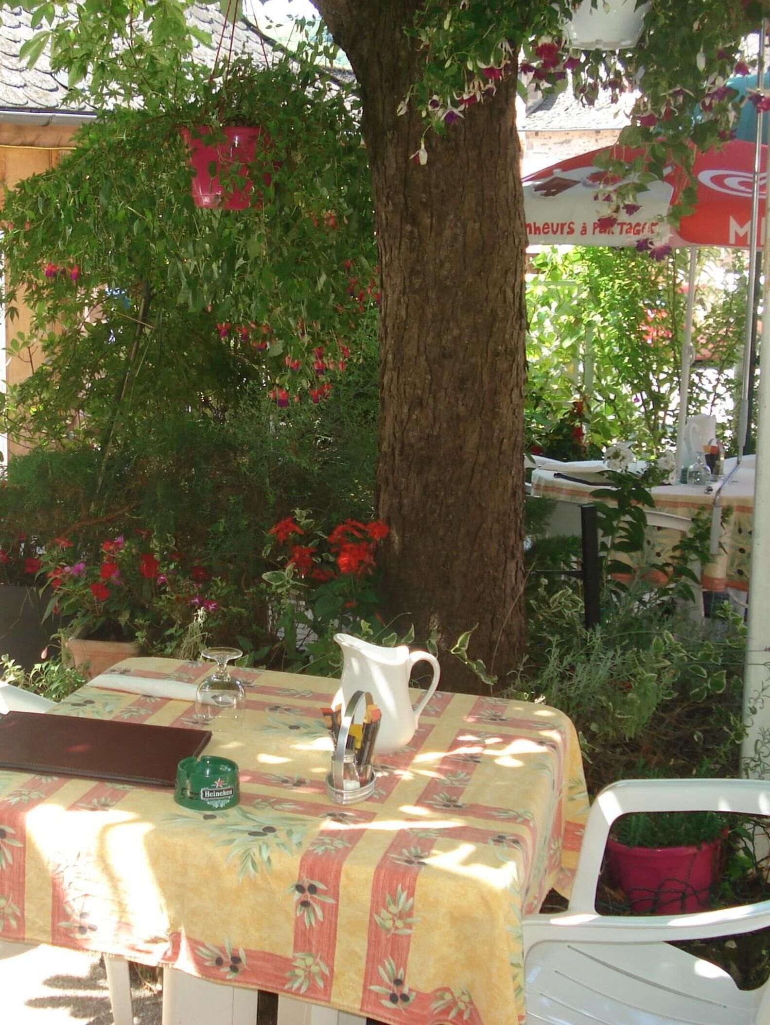 Uma área de estar externa com mesa coberta por toalha de mesa florida, um jarro branco e uma garrafa de Heineken.