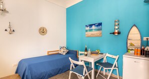 A room with a bed, a table with chairs, a clock, a painting of a beach, and a refrigerator. - Sea Apartment (Syracuse)