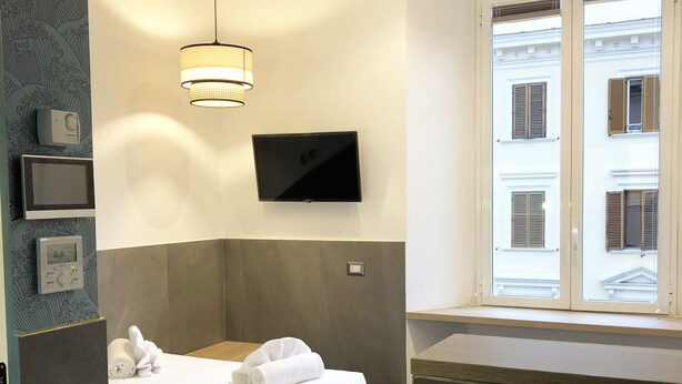 A modern hotel room with a bed, a flat-screen TV mounted on the wall, a window with shutters, and a wall-mounted control panel. - Ottaviano Smart Rooms (Rome)