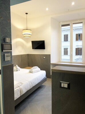 A modern hotel room with a bed, a flat-screen TV mounted on the wall, a window with shutters, and a wall-mounted control panel. - Ottaviano Smart Rooms (Rome)