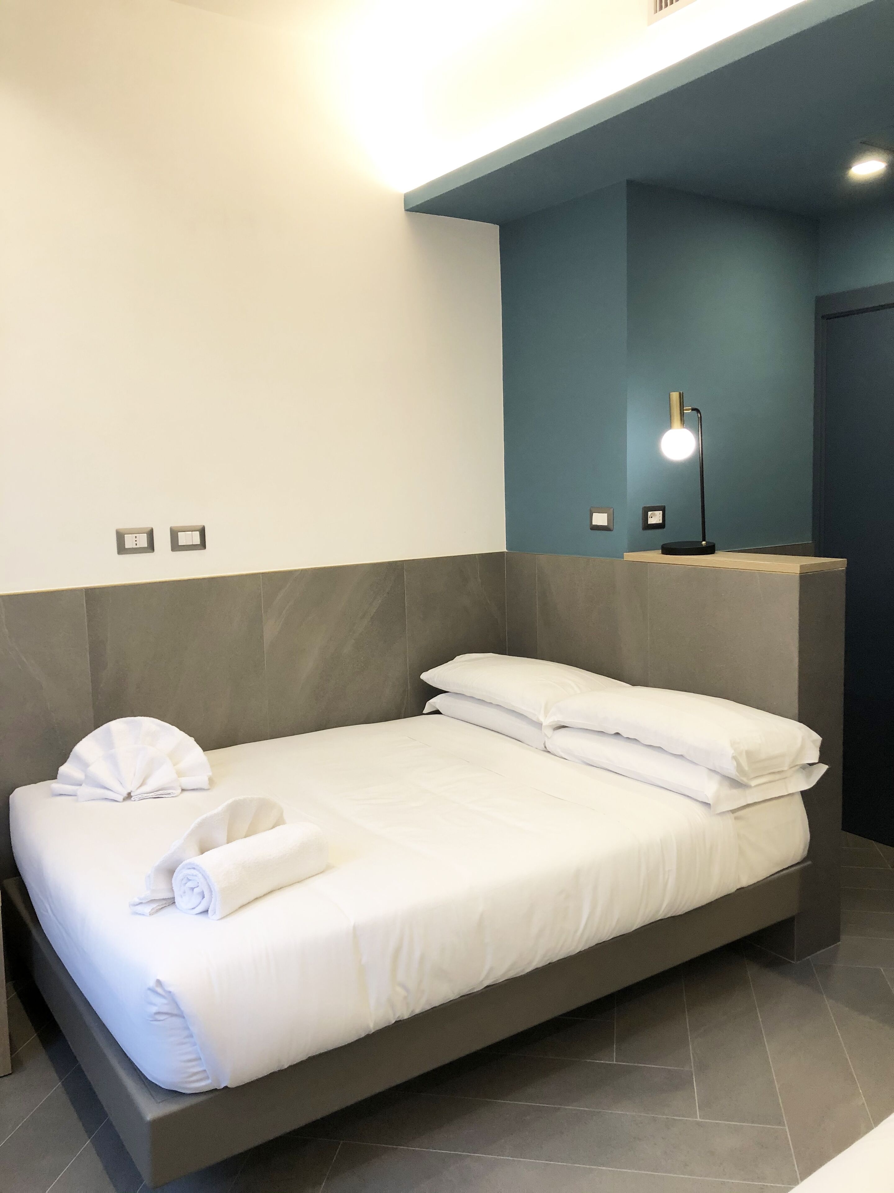 A modern hotel room with a bed, a nightstand, and a wall-mounted lamp.