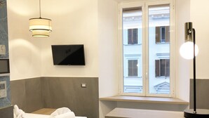A modern hotel room with a bed, a TV mounted on the wall, a window with a view of buildings, and a bedside table with a lamp. - Ottaviano Smart Rooms (Rome)