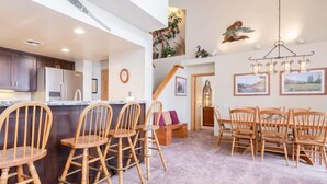A spacious kitchen with dark wooden cabinets, a stainless steel refrigerator, and a built-in island with bar stools. There is a dining area with wooden chairs and a chandelier. - Snowcreek #856, Updated 2 Bedrm + Loft Townhme in Snowcreek Phase 5 (Mammoth Lakes)