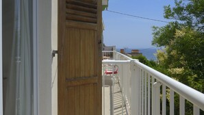 Uma varanda com porta de madeira, grades brancas e vista para o mar.