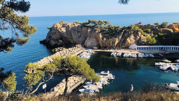 Uma marina costeira com barcos atracados, falésias rochosas e um edifício com a placa 'MAIDAGUE'.
