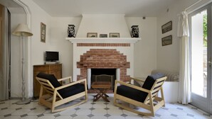 Uma sala de estar com lareira de tijolos, duas cadeiras de madeira com almofadas, uma televisão sobre um móvel e piso ladrilhado.
