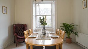 A dining room with a round wooden table set for six, surrounded by wooden chairs. A potted plant is on the right, and a red armchair is on the left. - Spacious 3BR New Town Apartment (Edinburgh)