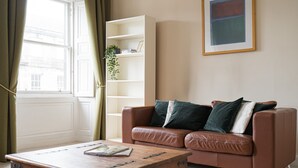 A living room with a brown leather sofa, a wooden coffee table, a white bookshelf, and a window with curtains. - Spacious 3BR New Town Apartment (Edinburgh)