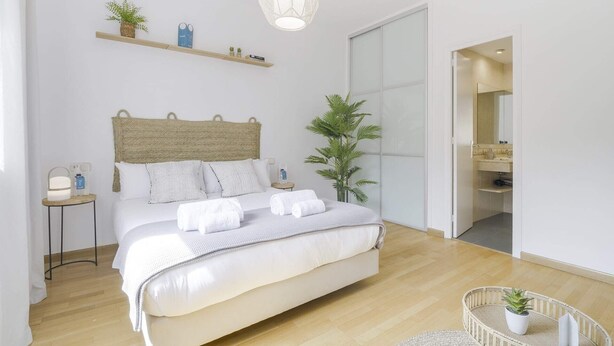 Un dormitorio moderno con una cama grande, una mesita de noche con lámpara, una lámpara colgante, un estante pequeño con objetos decorativos y una planta en maceta.