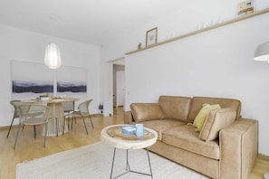 A modern living room with a beige sofa, a round coffee table, wicker chairs, a hanging light fixture, and a painting on the wall.