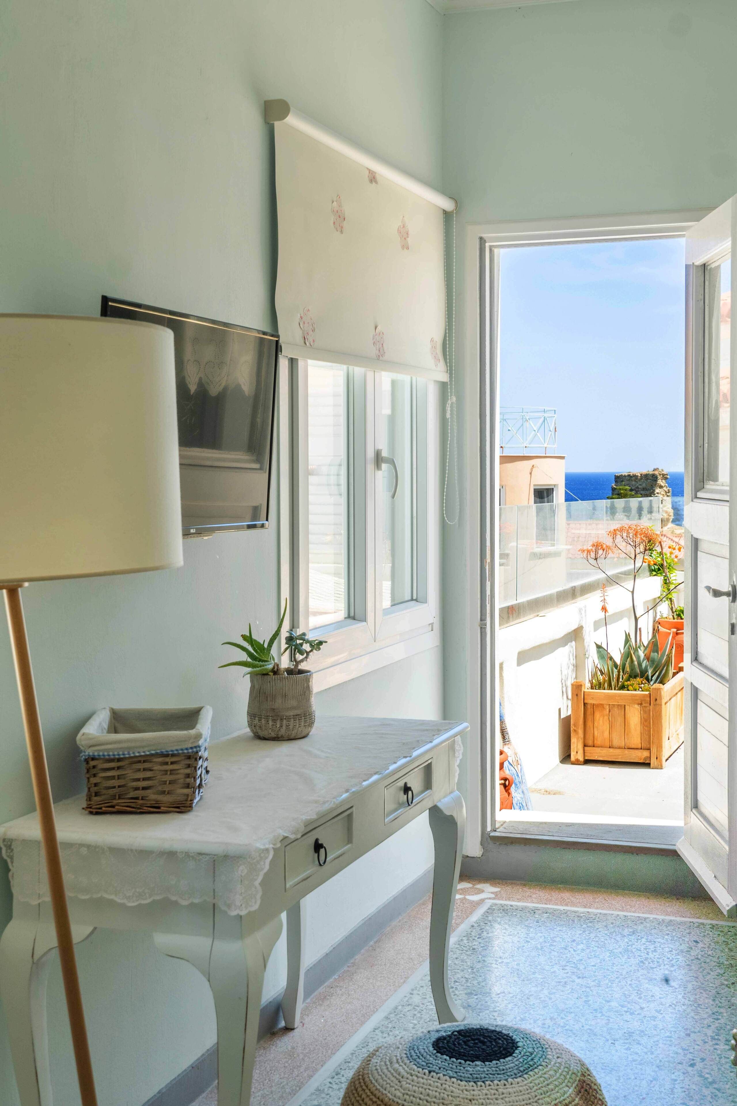 Una habitación con una mesa blanca, una lámpara, una planta en maceta y vistas al mar a través de una puerta abierta.