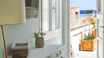 Una habitación con una mesa blanca, una lámpara, una planta en maceta y vistas al mar a través de una puerta abierta.