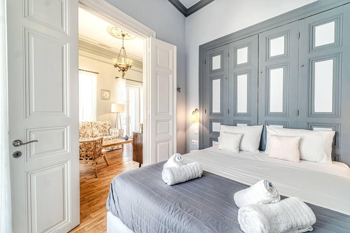 A hotel room with a large bed, a chandelier, a wooden floor, and a view of the hallway.