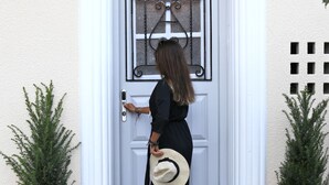 Uma mulher de vestido preto e chapéu bege em frente a uma porta branca com grade de janela decorativa. Há dois vasos de plantas de cada lado da porta.