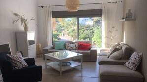 Ein Wohnzimmer mit Sofa, Sessel und Couchtisch, einem Schrank und Blick auf Grünflächen.