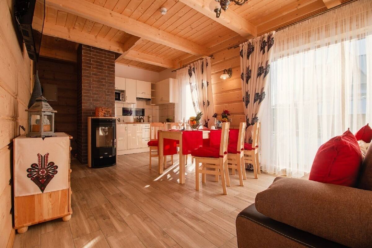 A cozy, well-lit living room with a fireplace, wooden floors, and a dining area with red chairs.
