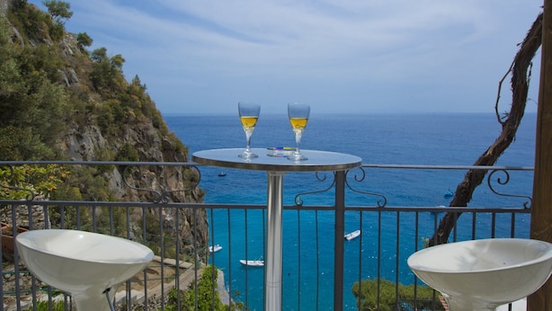 Uma varanda com duas cadeiras brancas, uma mesa redonda pequena e dois copos de vinho. A vista inclui um litoral rochoso e o mar.