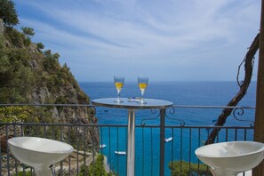 A balcony with two white chairs, a small round table, and two glasses of wine. The view includes a rocky coastline and the sea.