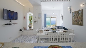 Uma sala de jantar com uma mesa branca posta para quatro pessoas, uma televisão na parede e vista para o mar por uma porta aberta.