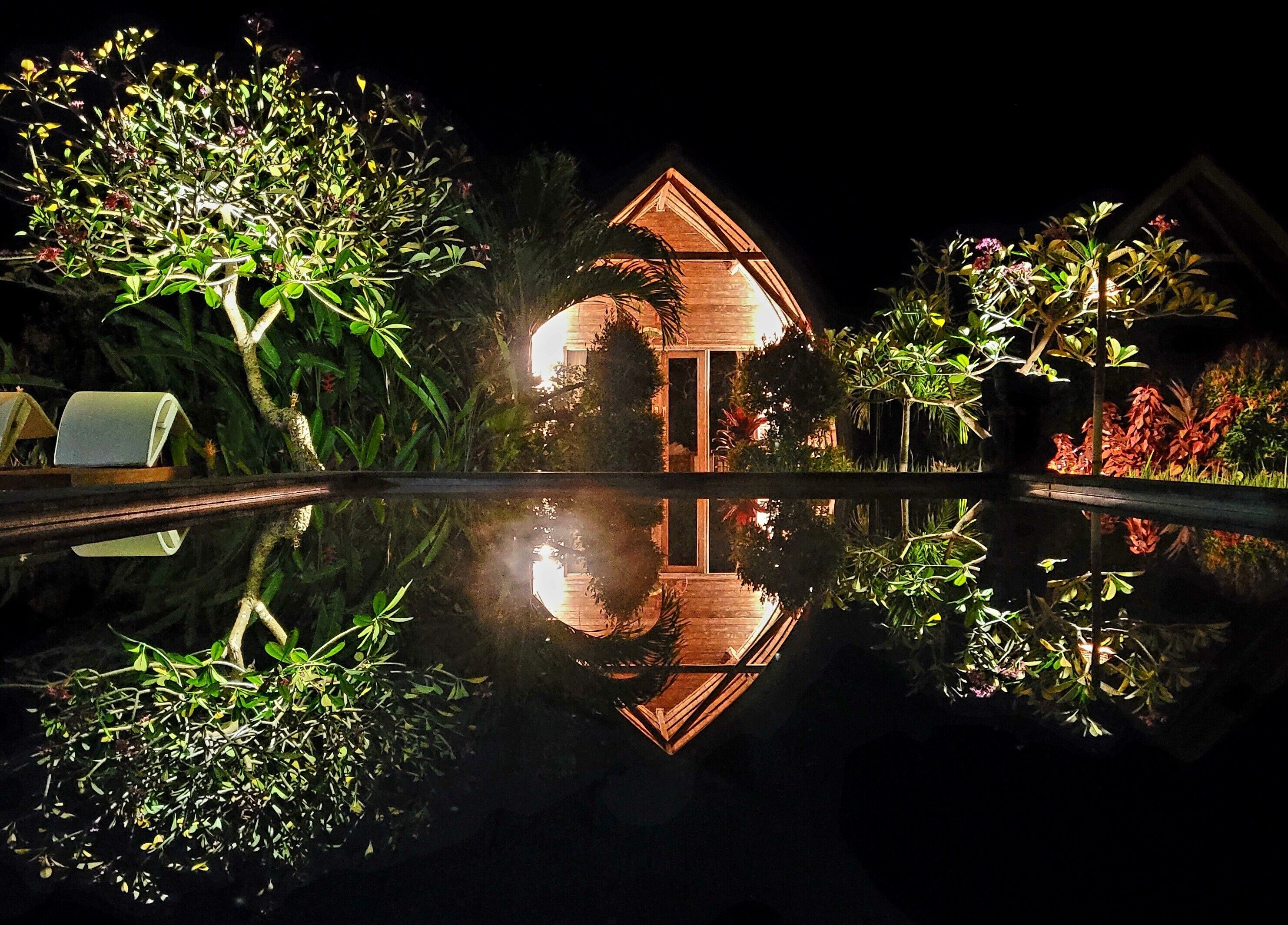 Une vue nocturne d’une maison dotée d’un toit triangulaire, entourée d’une végétation luxuriante et bordée d’un bassin dont la surface reflète la lumière.