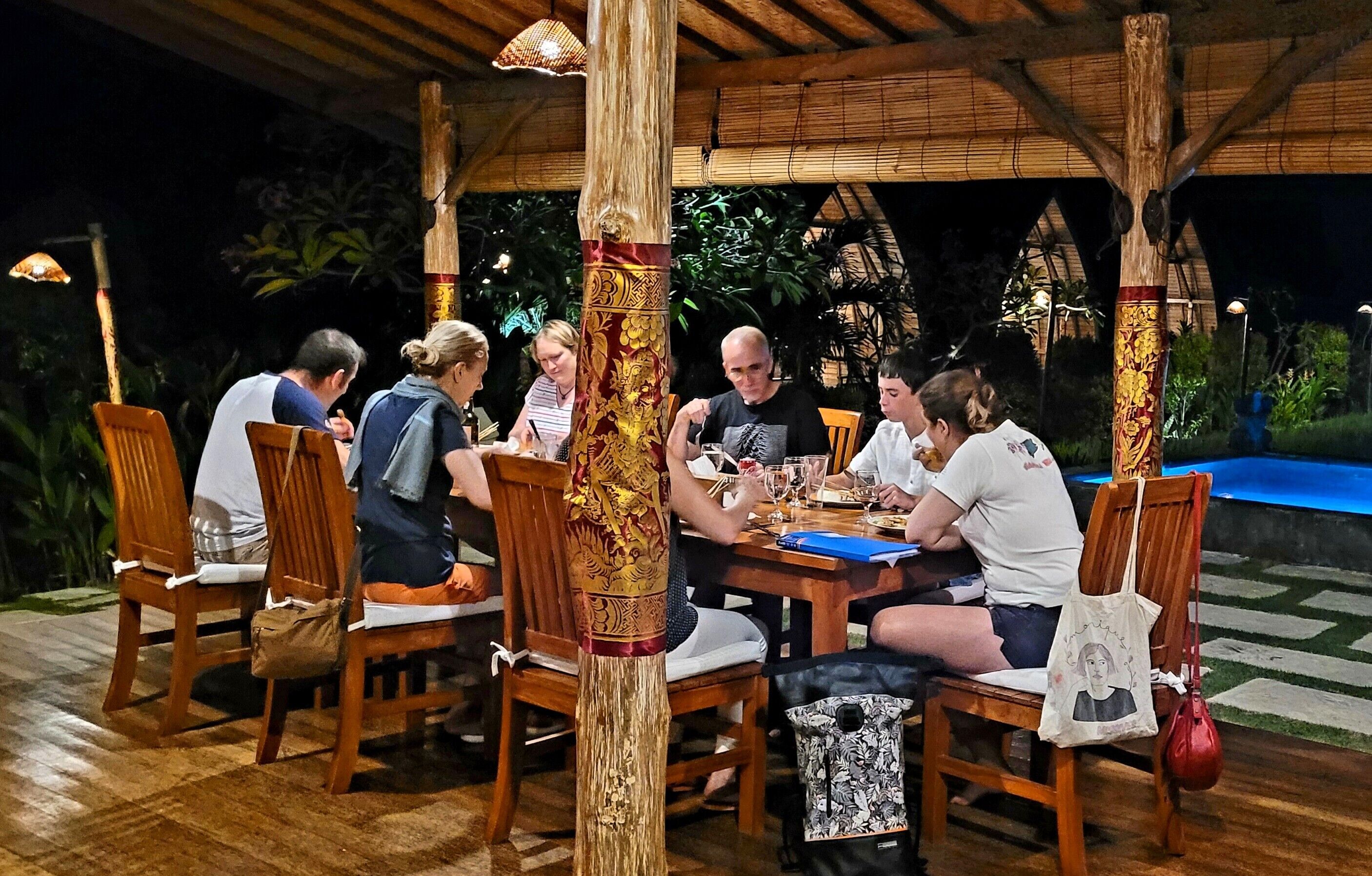 A group of people dining at an outdoor wooden table with chairs, surrounded by a bamboo structure and plants.