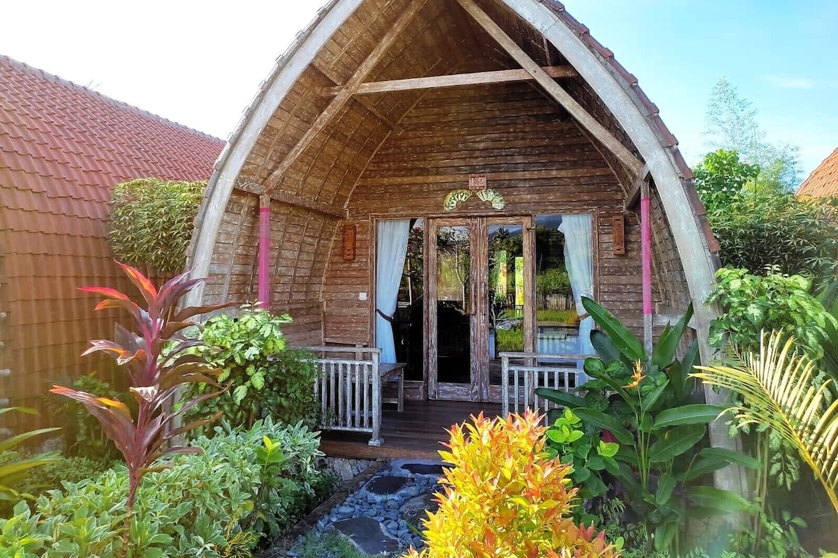 Un bâtiment rustique en bois, doté d’un toit à forte pente, d’un porche avec une balustrade et d’un petit jardin agrémenté de diverses plantes, ainsi que d’un chemin de pierre menant à l’entrée.