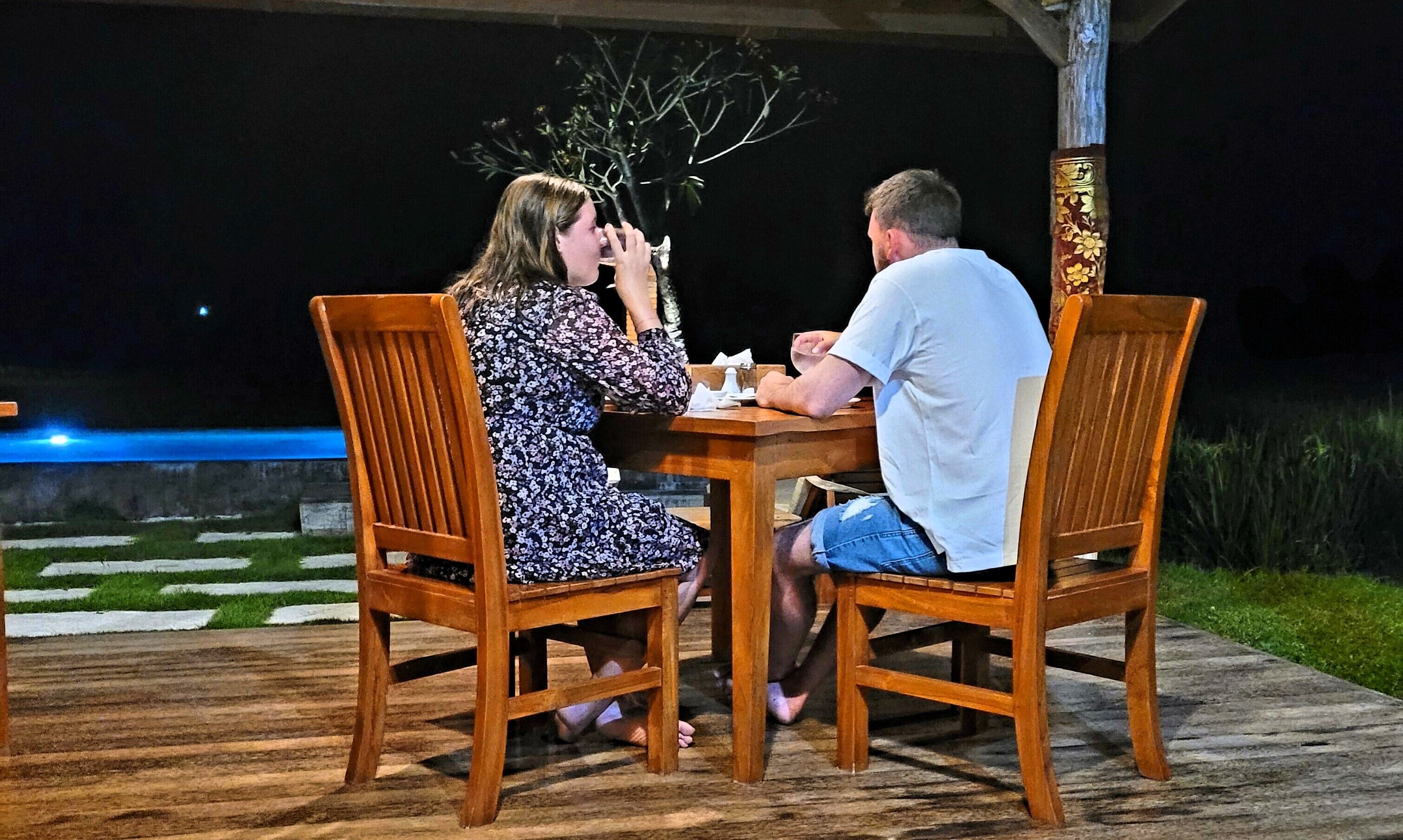Deux personnes sont assises à une table en bois à l’extérieur, de nuit. On aperçoit un arbre et un élément décoratif aquatique en arrière-plan.