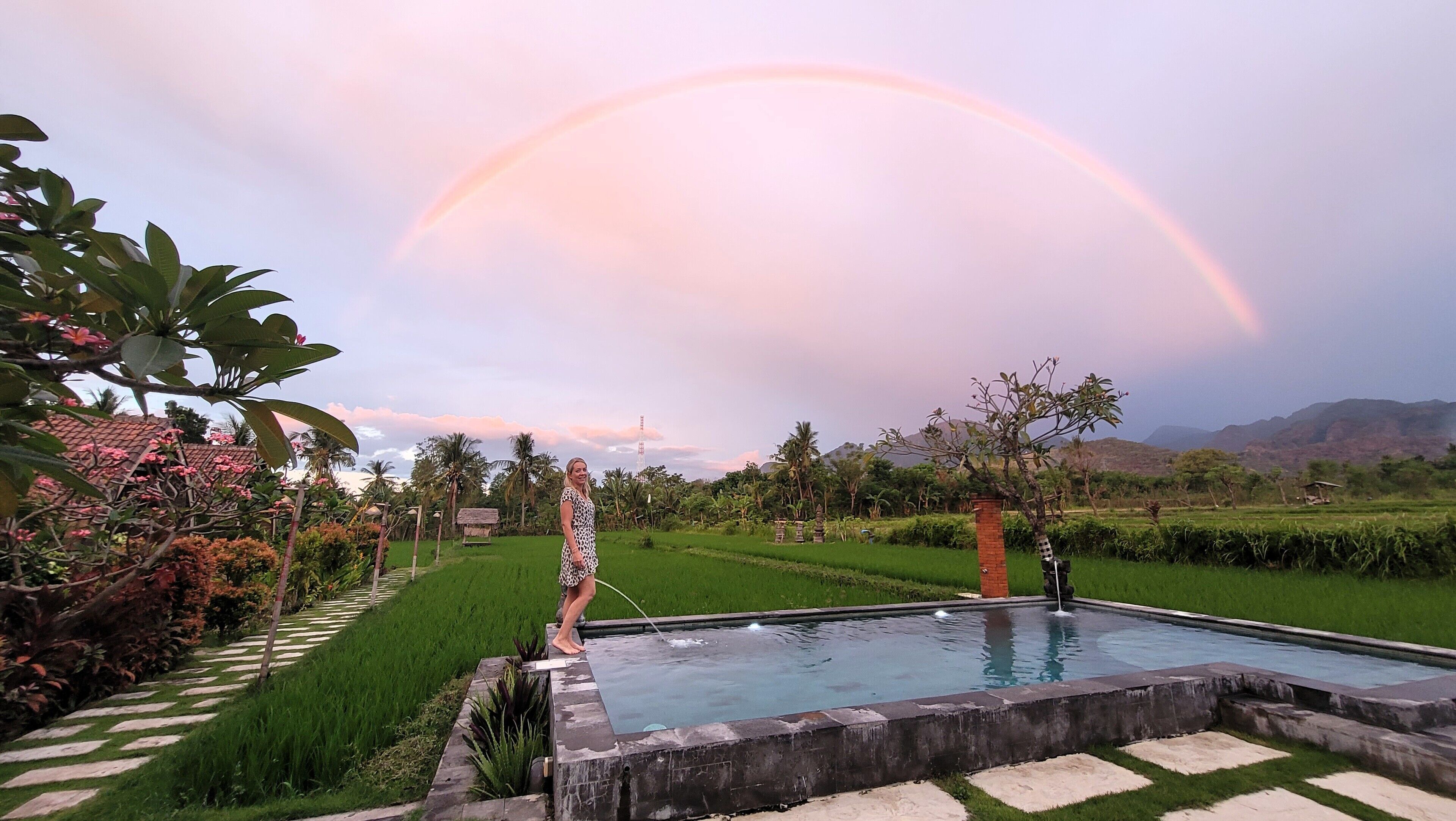Una piscina con borde de piedra, rodeada de vegetación exuberante y un cielo despejado con un arcoíris.