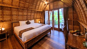 A rustic wooden room with a large bed, a wooden desk, and a balcony with a view of greenery. - Umma Bali Menjangan (Pejarakan)