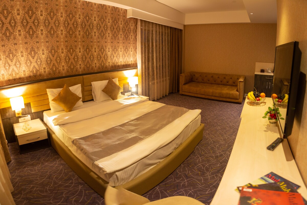 A hotel room with a large bed, a sofa, a desk with a mirror, and a TV.