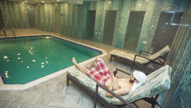 Une piscine avec deux chaises longues, dont l’une est occupée par une personne portant un short à carreaux et tenant une serviette, et l’autre est vide.