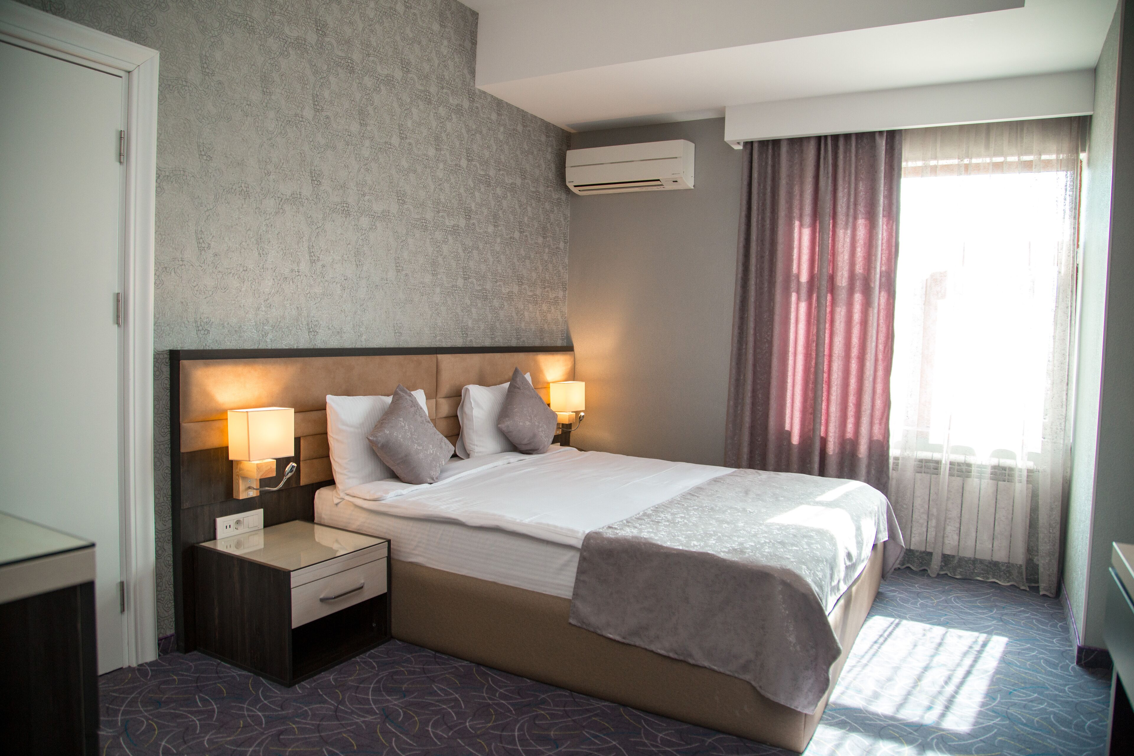 A hotel room with a large bed, two bedside tables with lamps, a headboard, and a window with curtains.