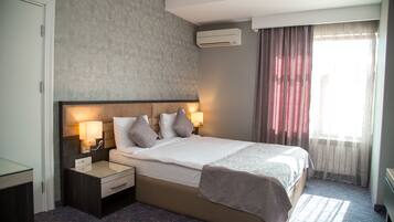 Una habitación de hotel con una cama grande, dos mesitas de noche con lámparas, un cabecero y un ventanal con cortinas.