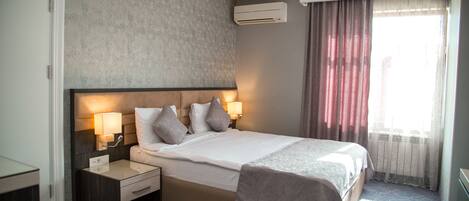 A hotel room with a large bed, two bedside tables with lamps, a headboard, and a window with curtains.