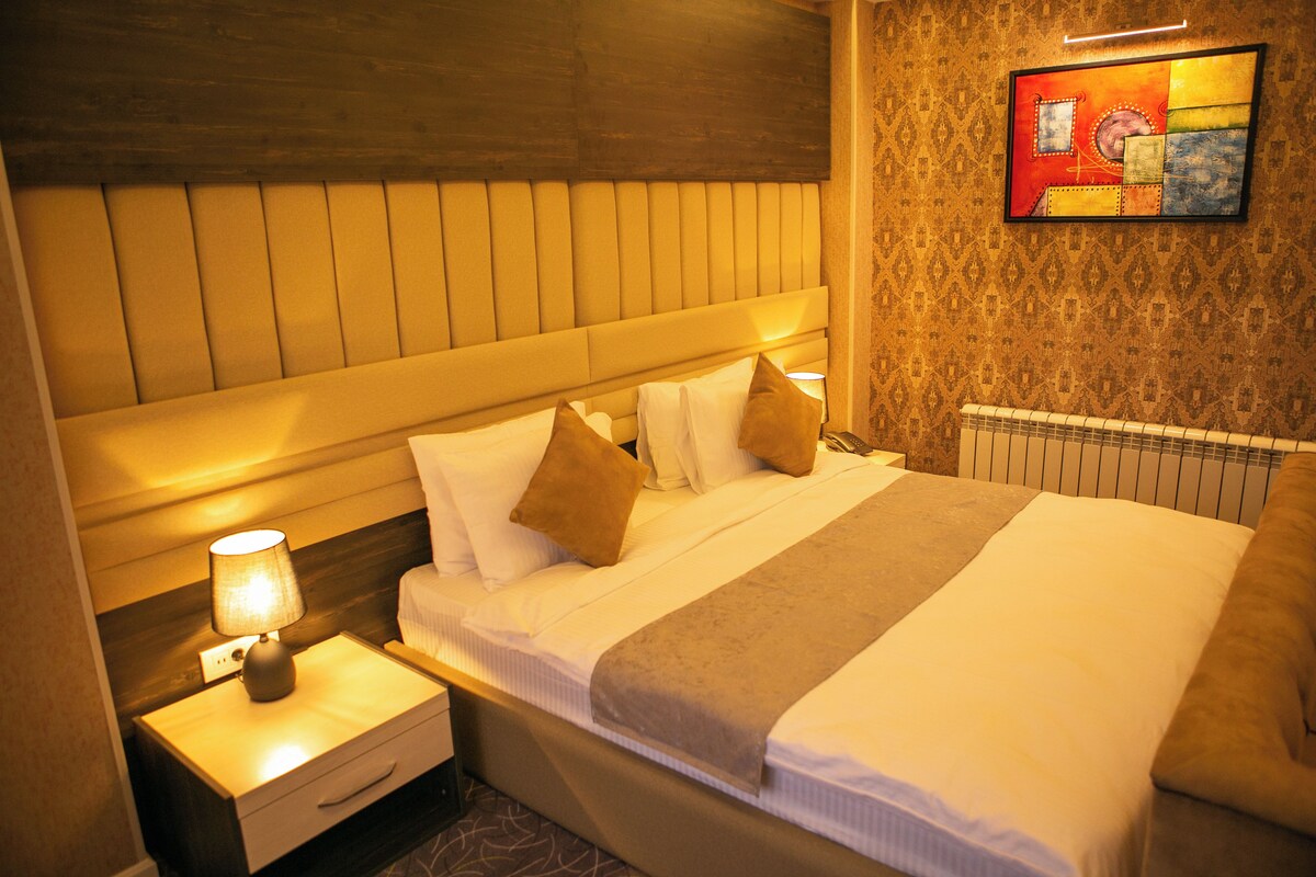 A hotel room with a large bed, a nightstand with a lamp, a wall-mounted painting, and a patterned wall.