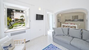 Una sala de estar moderna con un sofá gris, un televisor montado en la pared y vistas a un jardín a través de un ventanal.