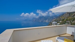 Un balcón con vistas al mar y a las montañas, una sombrilla blanca y dos sillones reclinables a rayas.