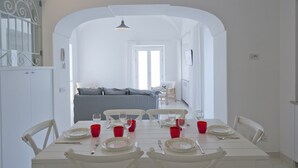 Un comedor con una mesa blanca dispuesta para cuatro, tazas rojas y vistas a una sala de estar con un sofá y una silla.